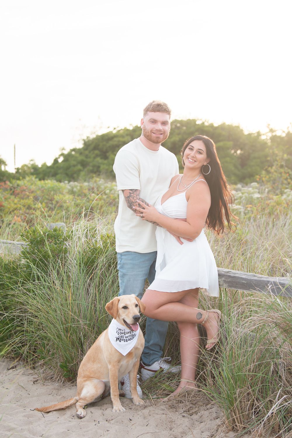 A couple poses with their yellow lab dog wearing a bandana on a sandy beach path at sunset.