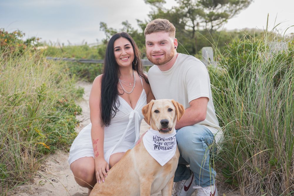 A couple kneels with their yellow lab dog wearing a white bandana among beach grass.