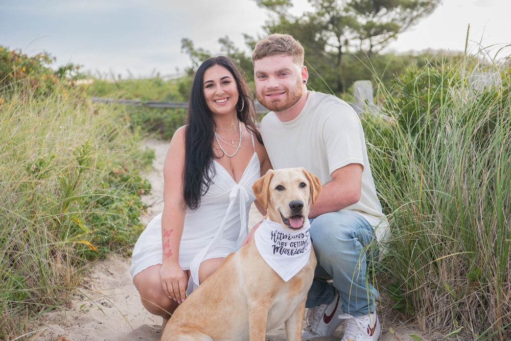 A couple and their yellow lab dog pose together on a sandy path surrounded by beach grass.