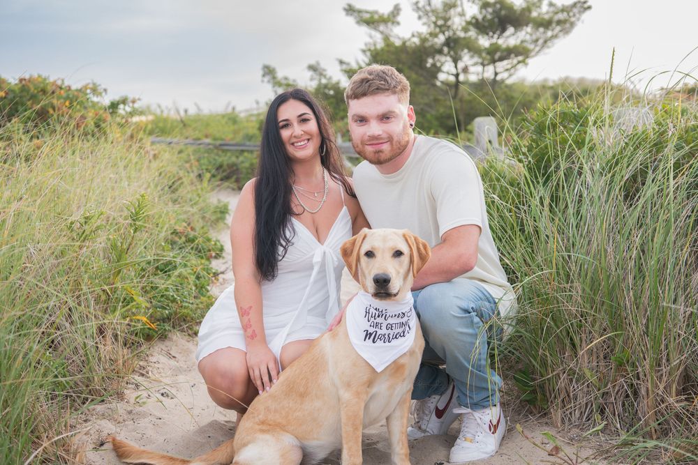 A couple poses with their yellow lab dog on a sandy path with beach grass in the background.