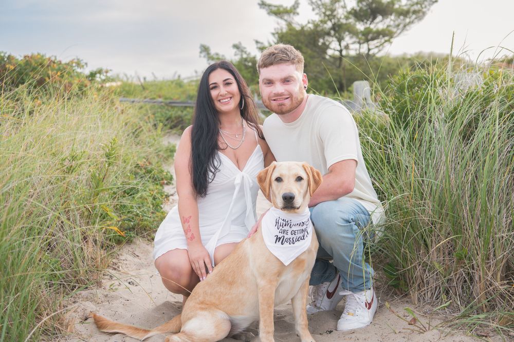 A couple smiles with their yellow lab wearing a bandana while kneeling on a beach path.