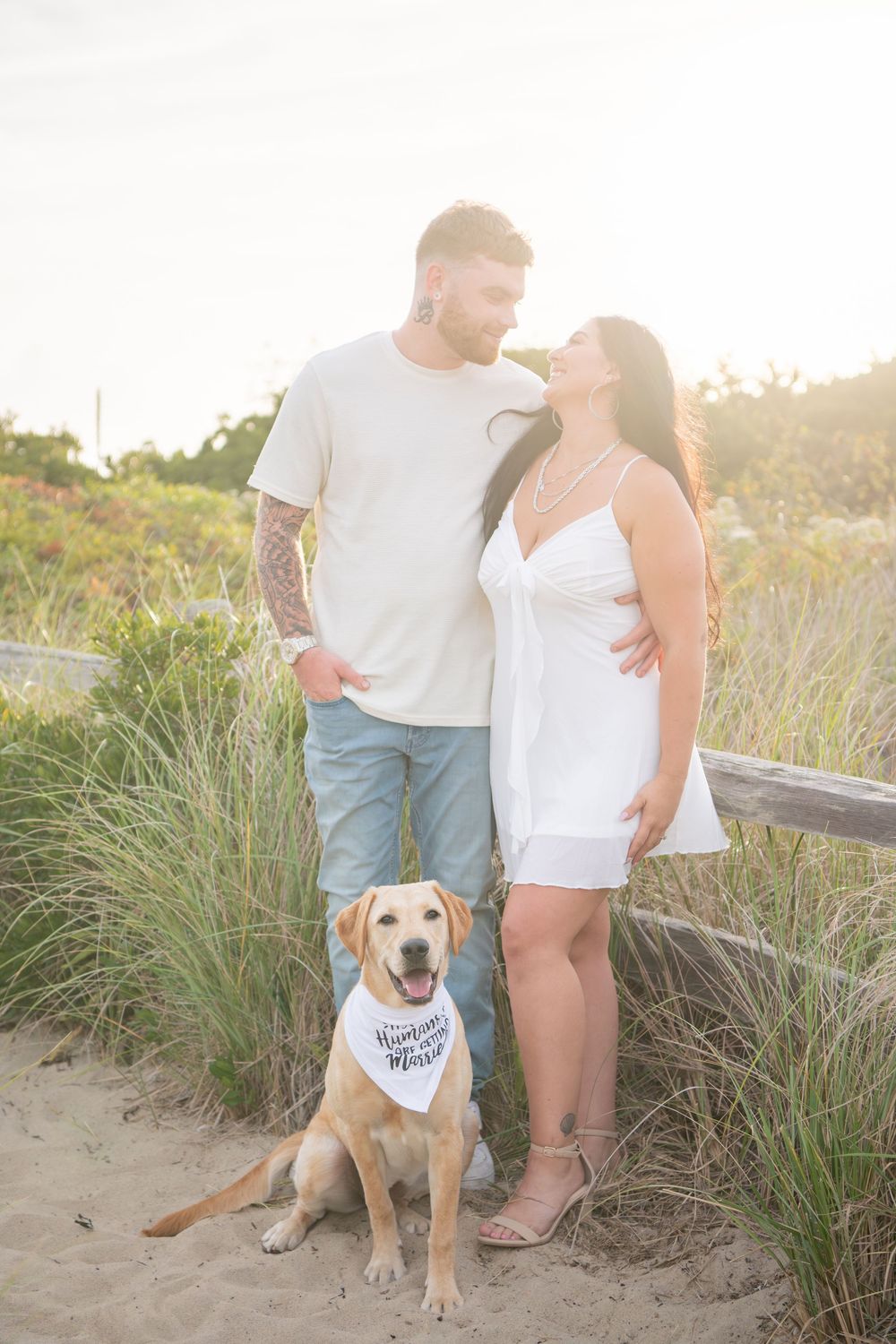 Couple with their yellow lab dog wearing bandana on beach path at sunset.