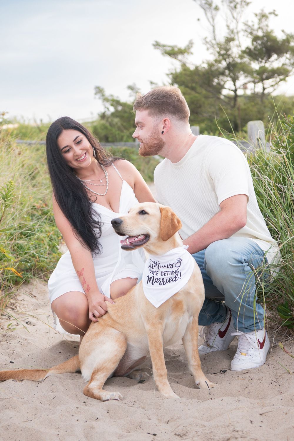 A couple laughs with their yellow lab dog wearing a 'She Said Yes' bandana on a beach path.