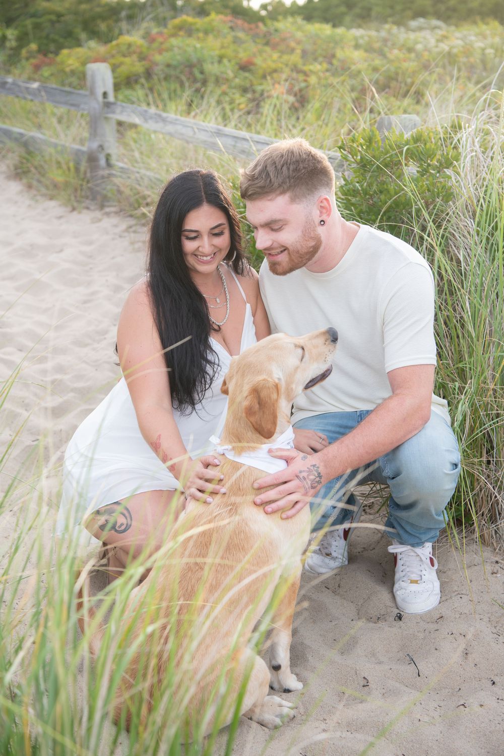 A couple pets their yellow lab dog while sitting on a sandy beach path surrounded by grass.