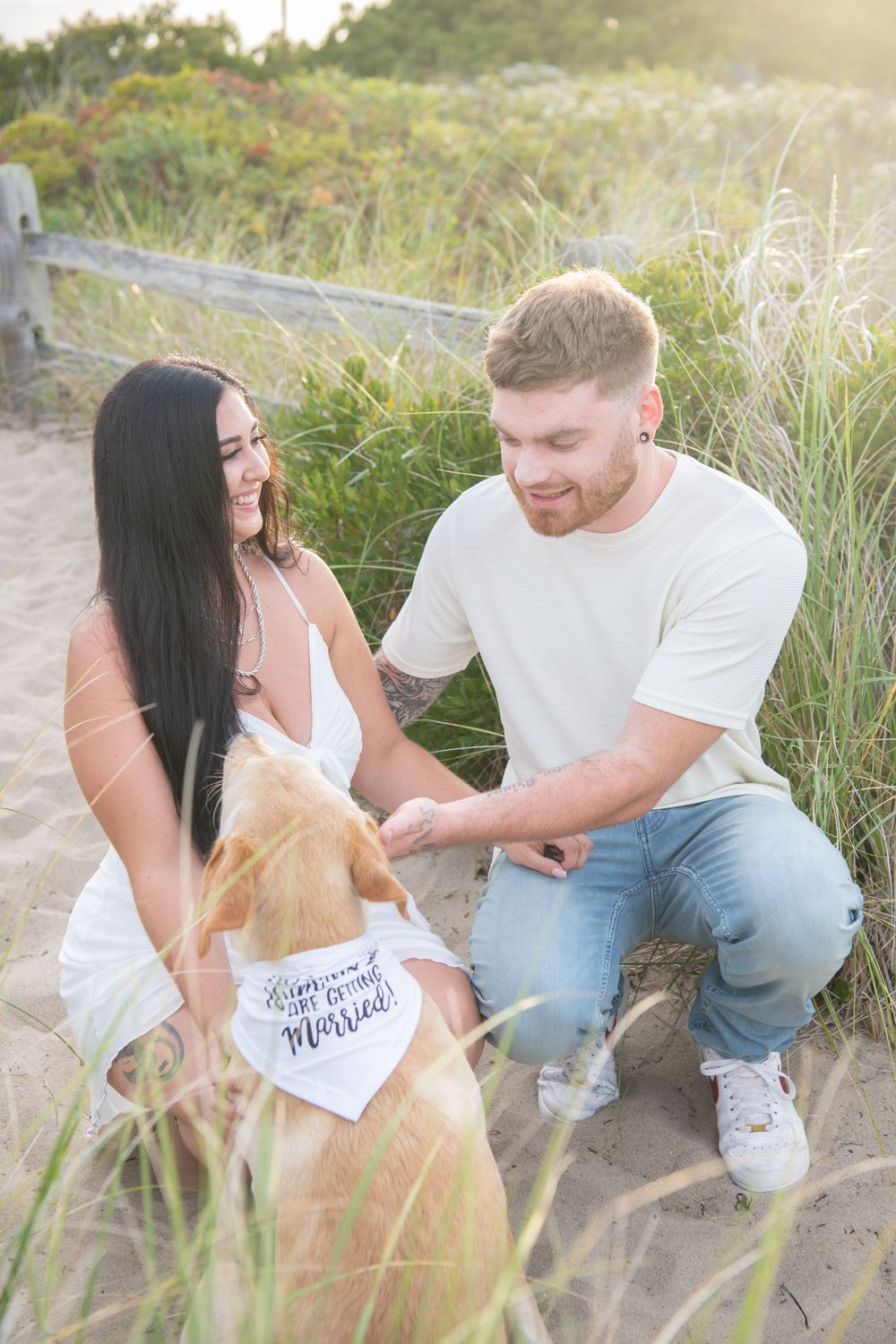 A couple shares a joyful moment with their yellow lab dog wearing a white bandana on a beach path.