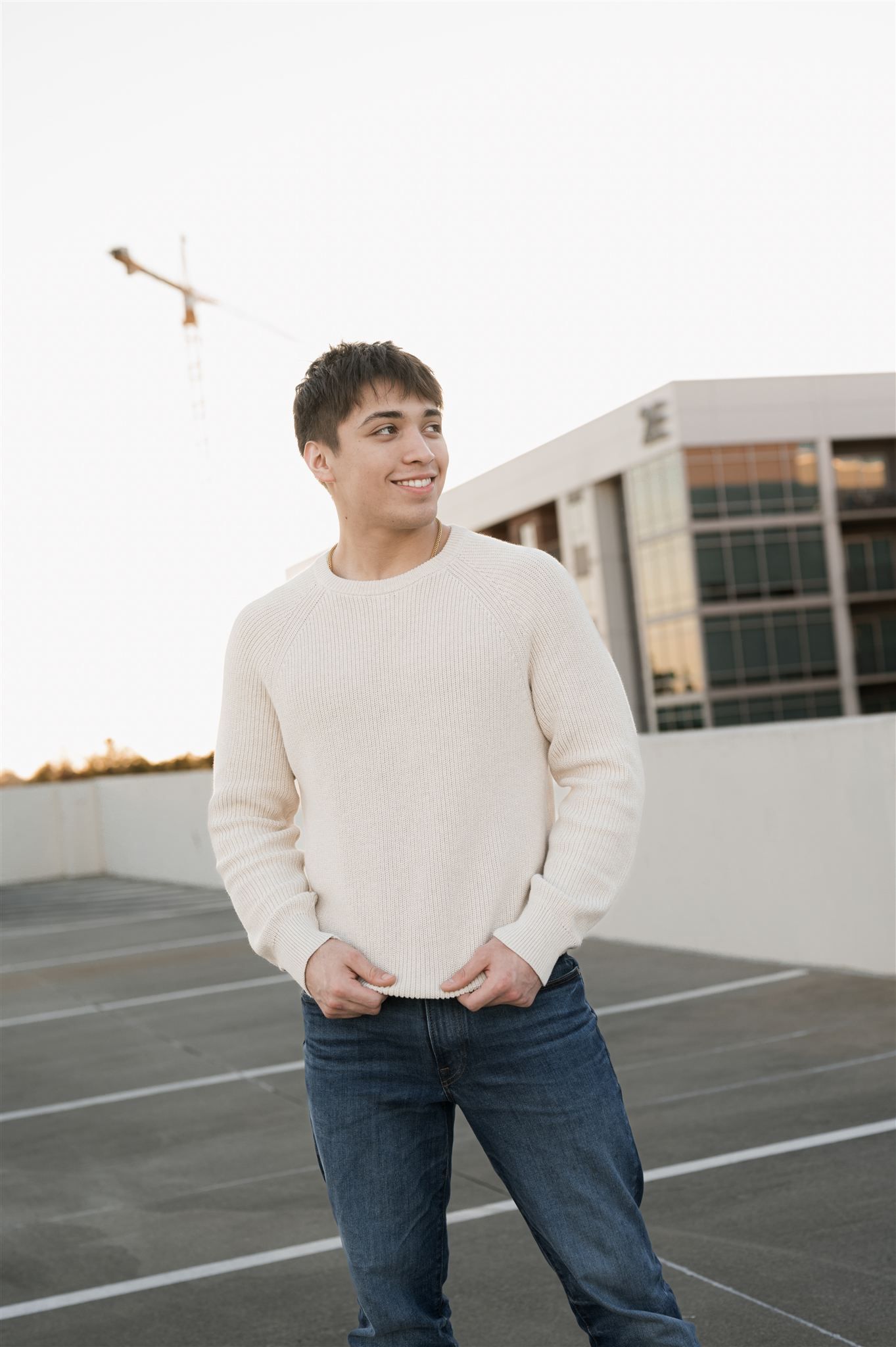 Person in a cream sweater and blue jeans stands in an empty parking lot at sunset.