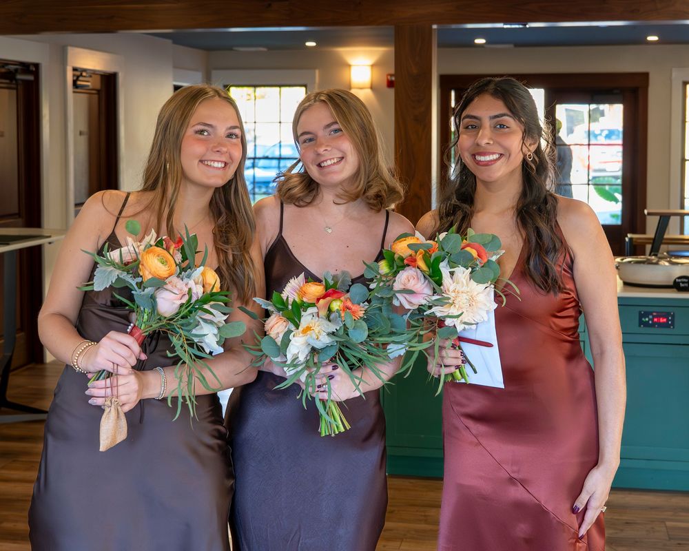 Three bridesmaids in brown and mauve dresses hold colorful floral bouquets with orange and white flowers indoors.