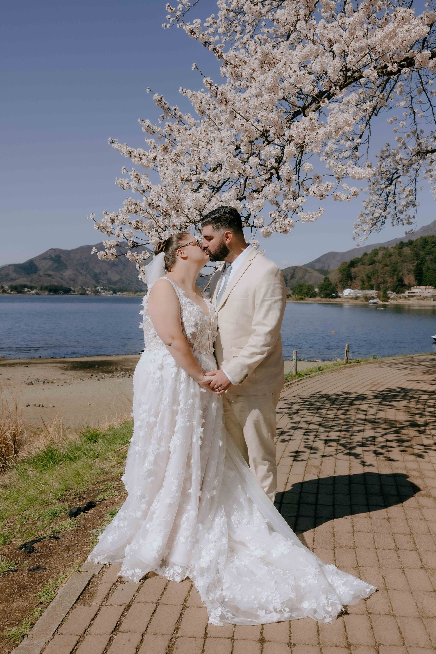 A romantic wedding couple embraces beneath blooming cherry blossoms by a scenic lake with mountains in the background.