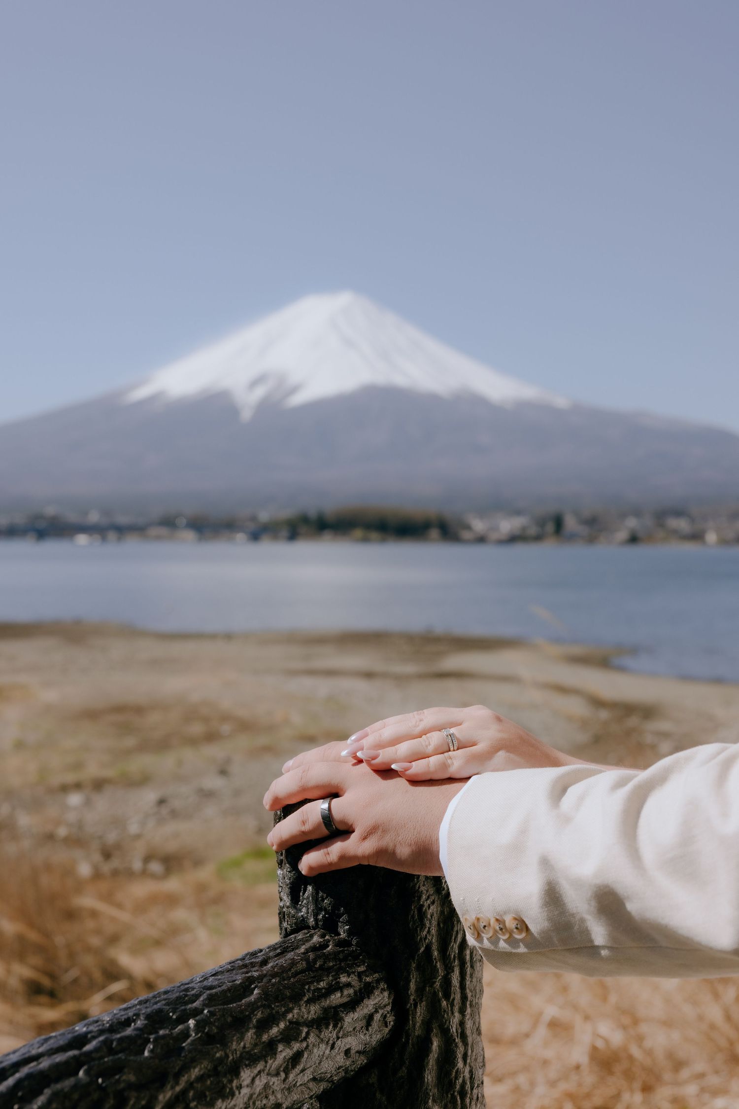 Hand rests on dark rock formation with snow-capped Mount Fuji rising majestically in the distance across a lake.