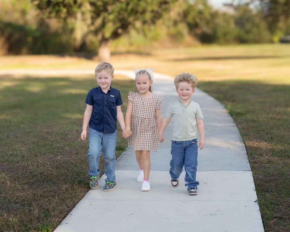 Three children walk and play together on a concrete path through a grassy field on a sunny day.