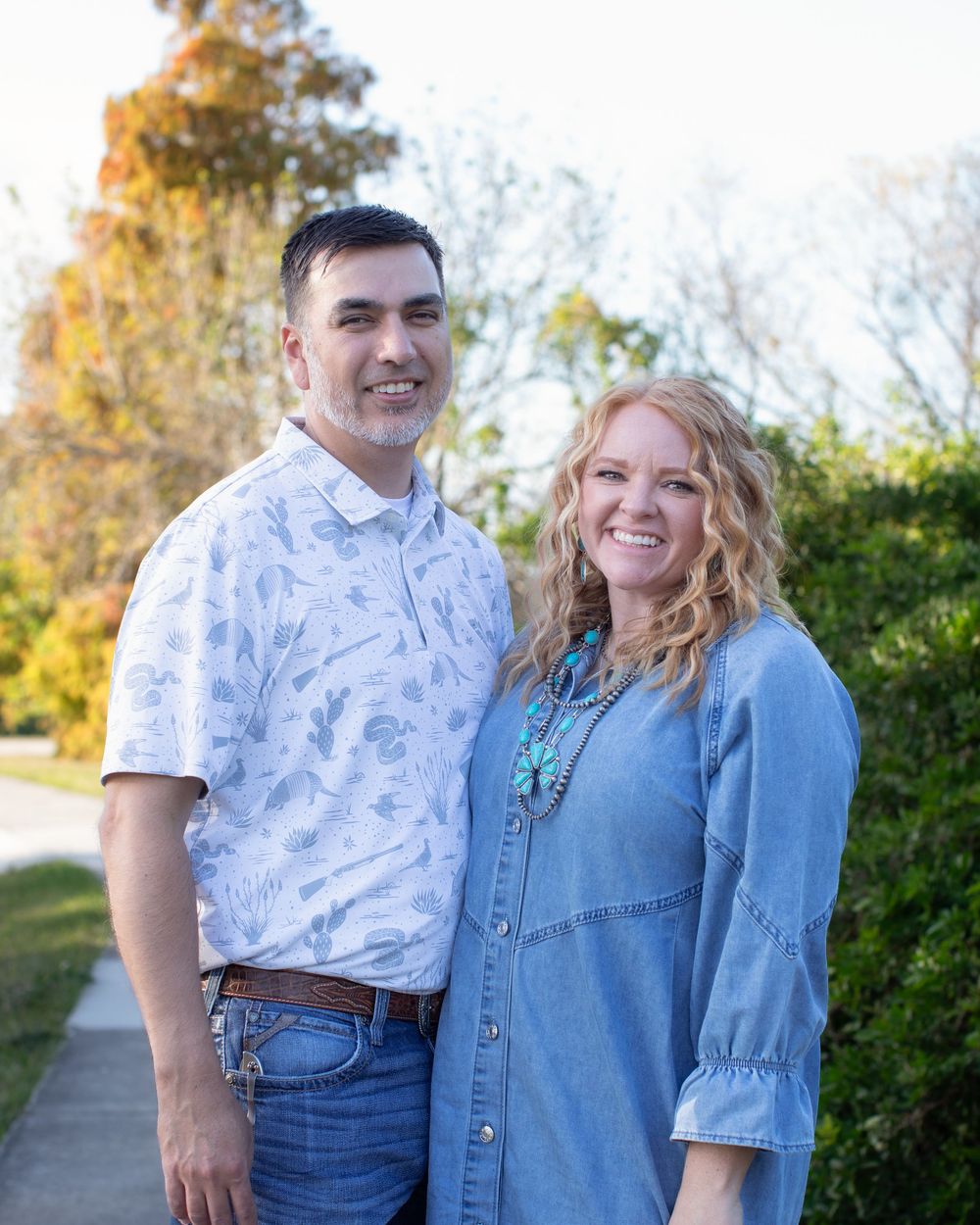 A couple poses together outdoors wearing coordinating denim and casual attire against a natural backdrop.