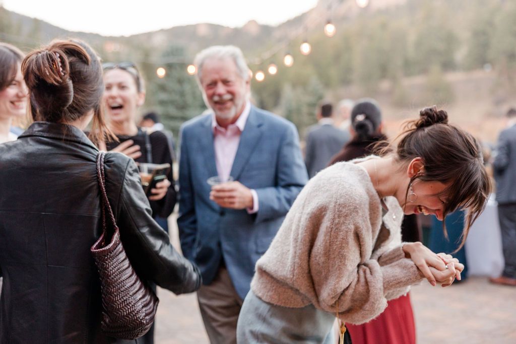 Wedding guests gather outdoors at a rustic venue with pine trees and string lights creating a festive atmosphere.