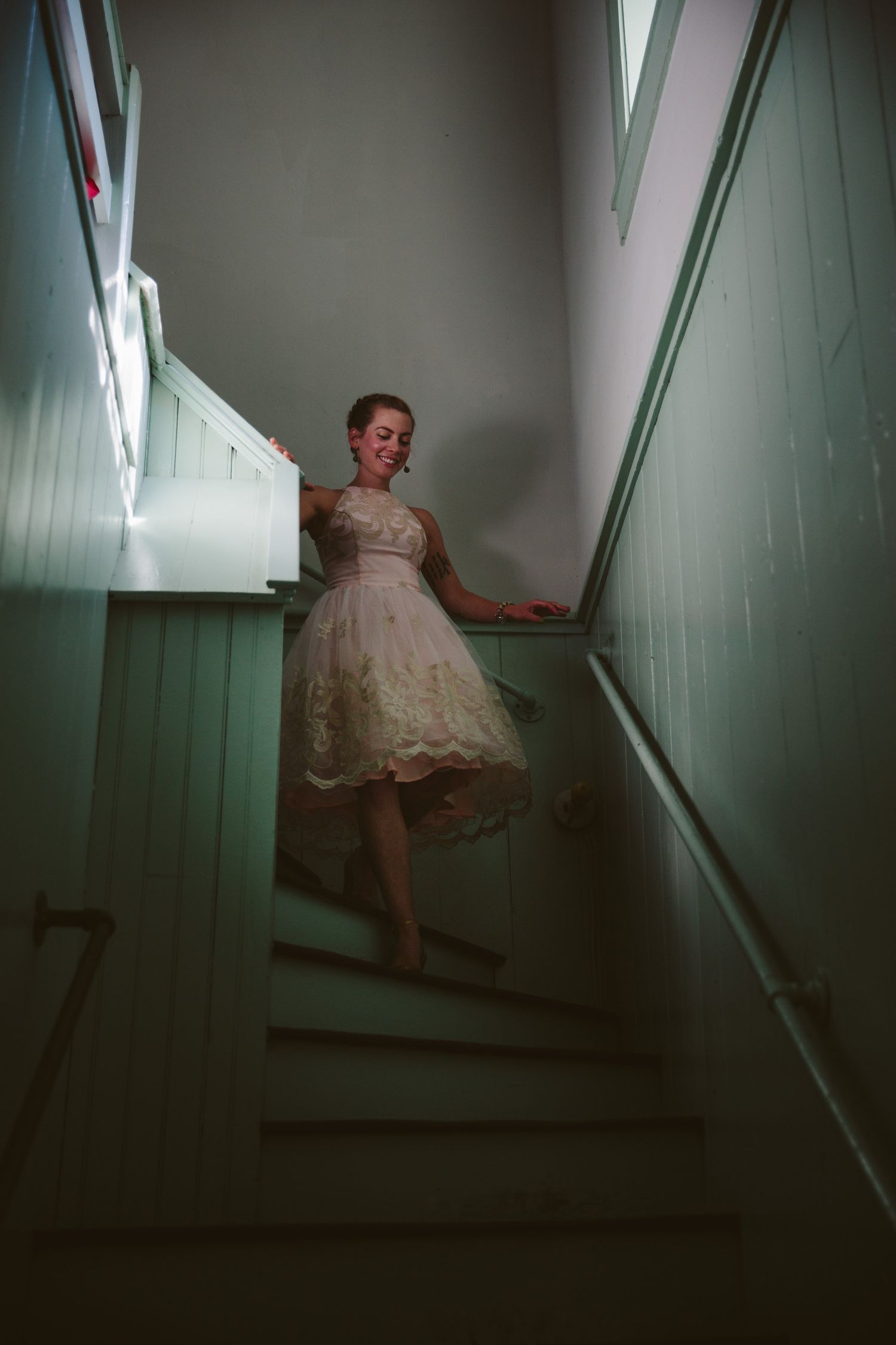 A figure in a vintage white lace dress stands on dimly lit mint green stairs in a moody, atmospheric setting.