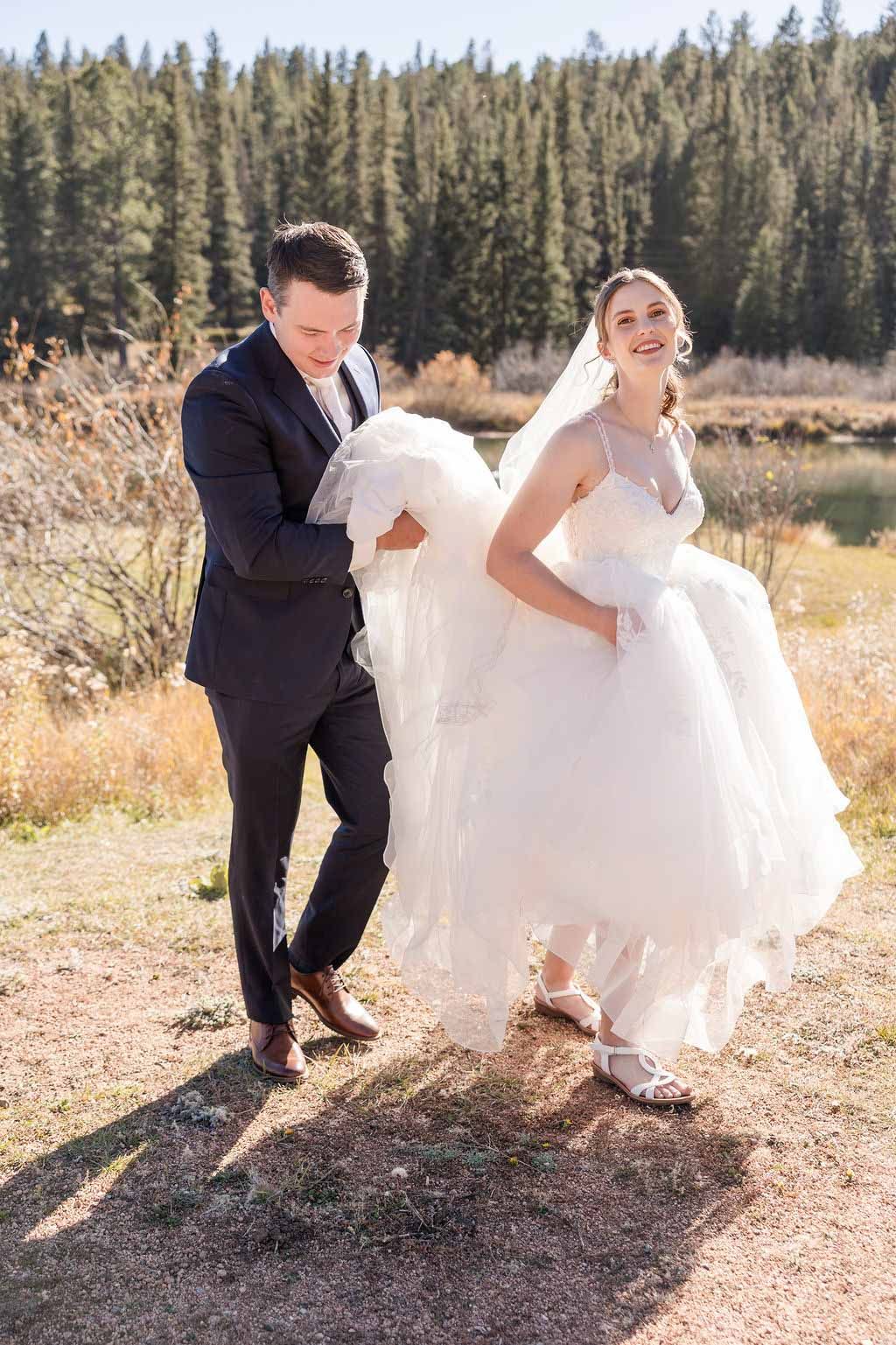 Playful wedding couple runs through golden field with pine trees in background.