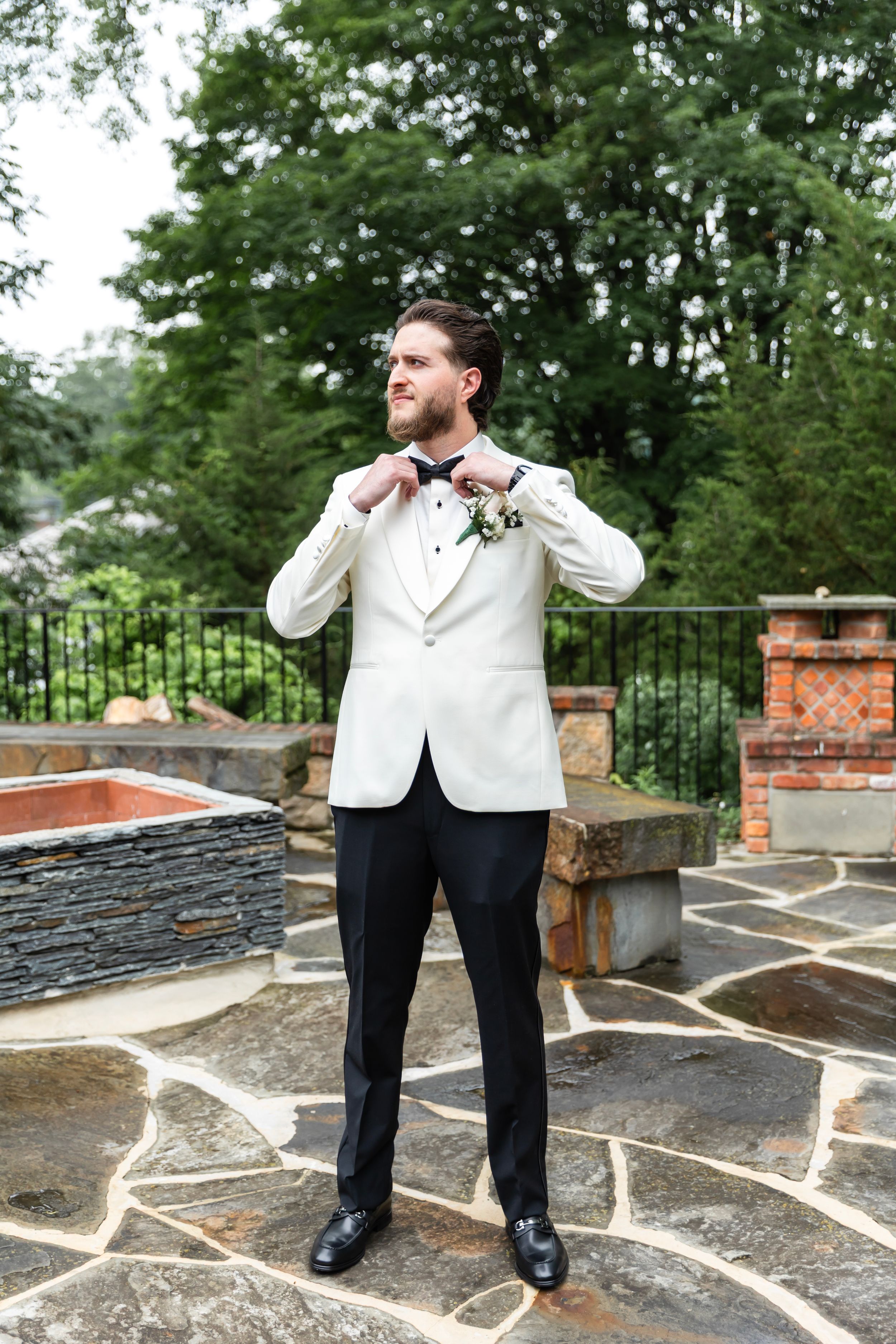 Groom in a white tuxedo jacket and black pants stands on a stone patio while talking on a phone. Sussex County wedding photographer