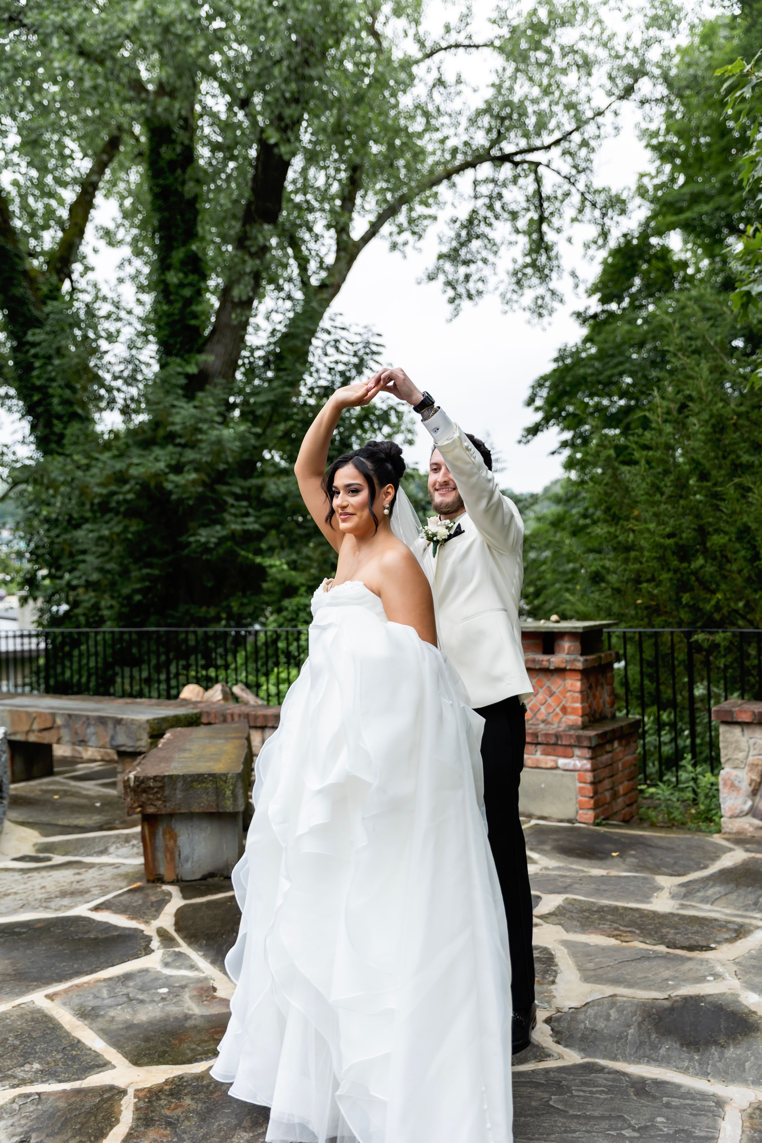 A couple in wedding attire dances together on a stone terrace surrounded by trees and brick pillars. Sussex County wedding photographer
