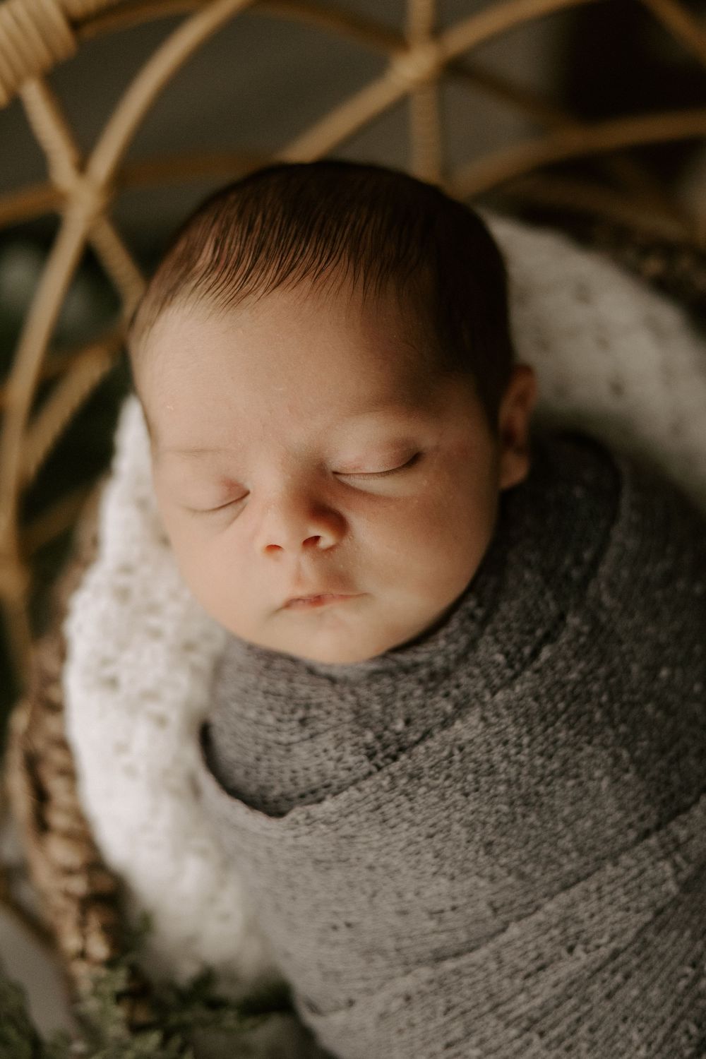 A sleeping newborn baby wrapped snugly in a soft gray knit blanket against a neutral backdrop.