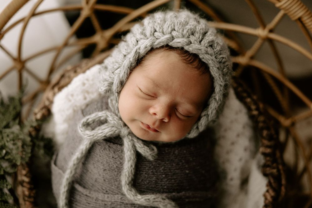 A sleeping baby wrapped in soft gray swaddle blankets lies peacefully in a rustic wooden basket.