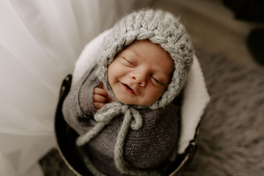 A sleeping newborn wrapped in a cozy gray knit bonnet and blanket rests peacefully on white fabric.