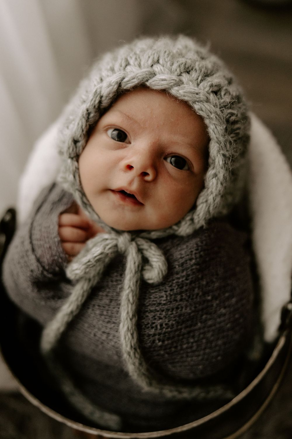 A baby wrapped in a cozy gray knit blanket and hat looks up with bright eyes and rosy cheeks.