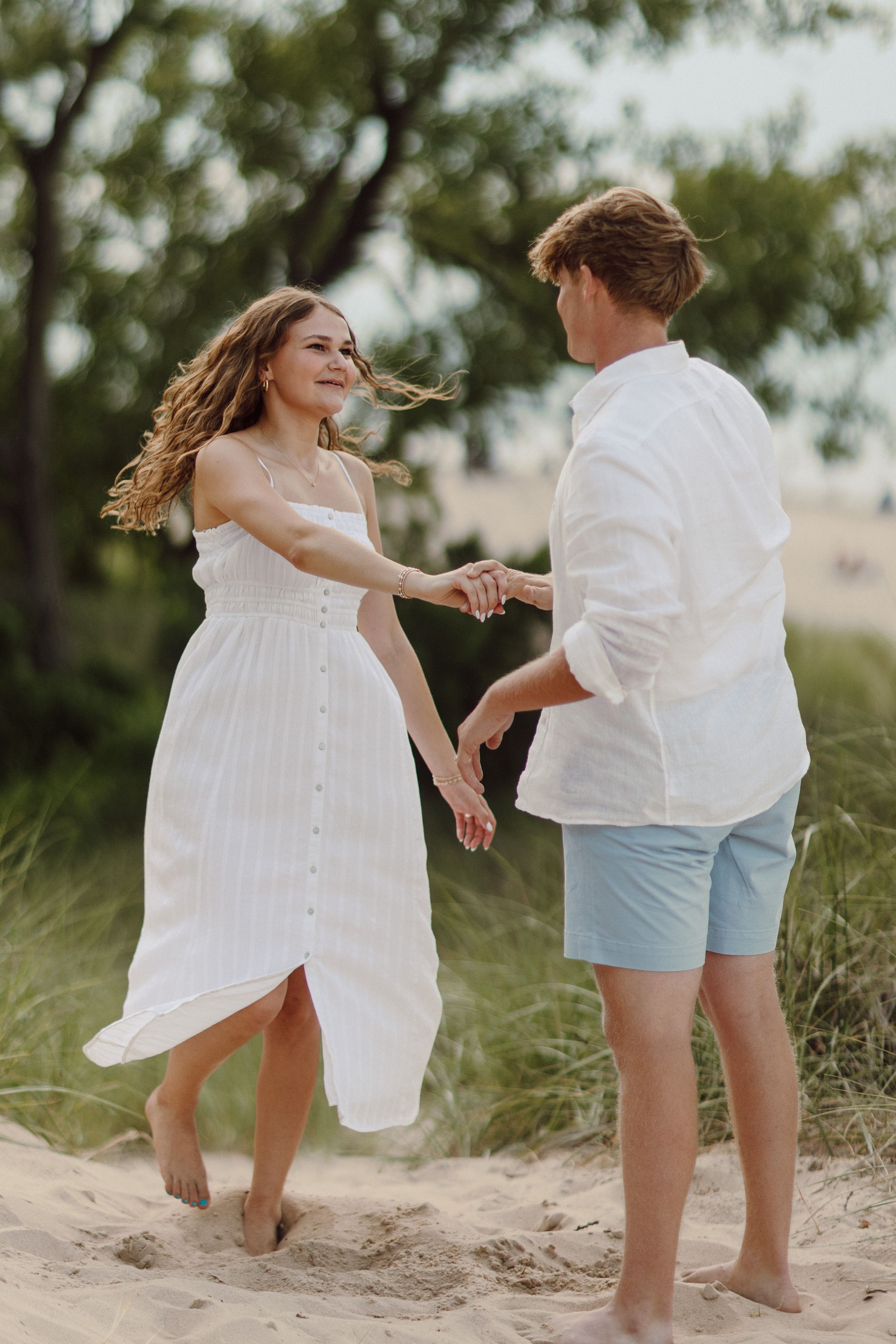 Playful couple in white clothing dancing on sandy beach dunes.