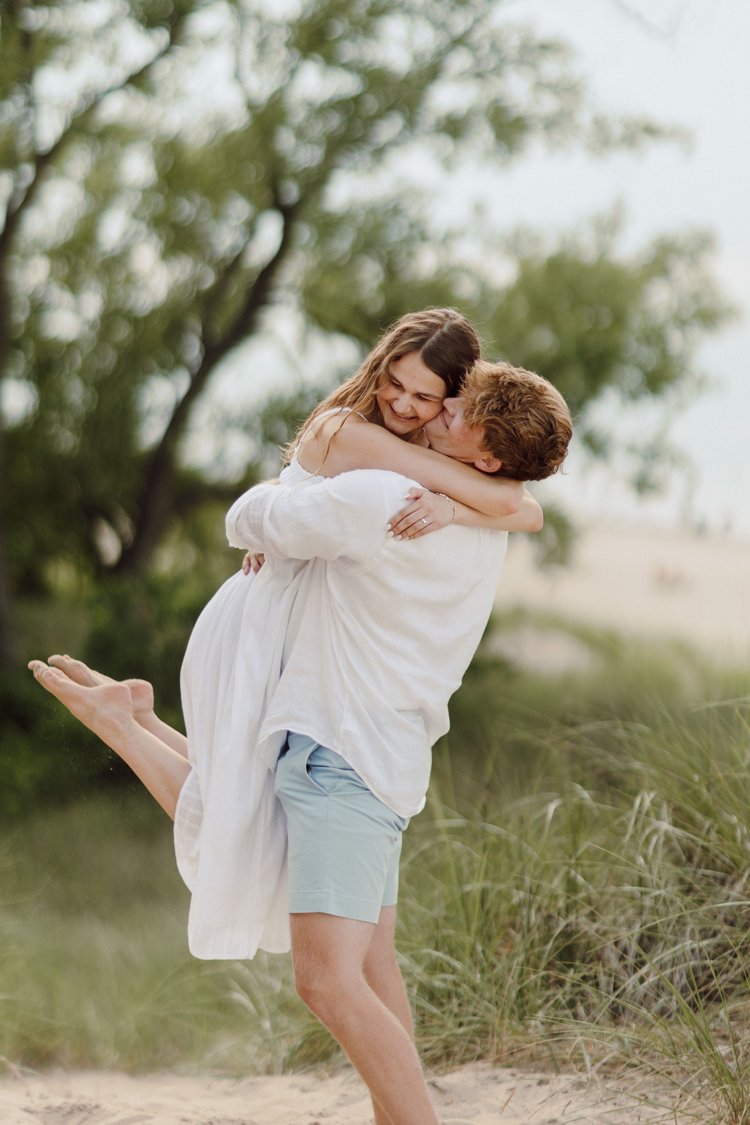 A romantic couple embraces on a sandy beach with green foliage in the background.