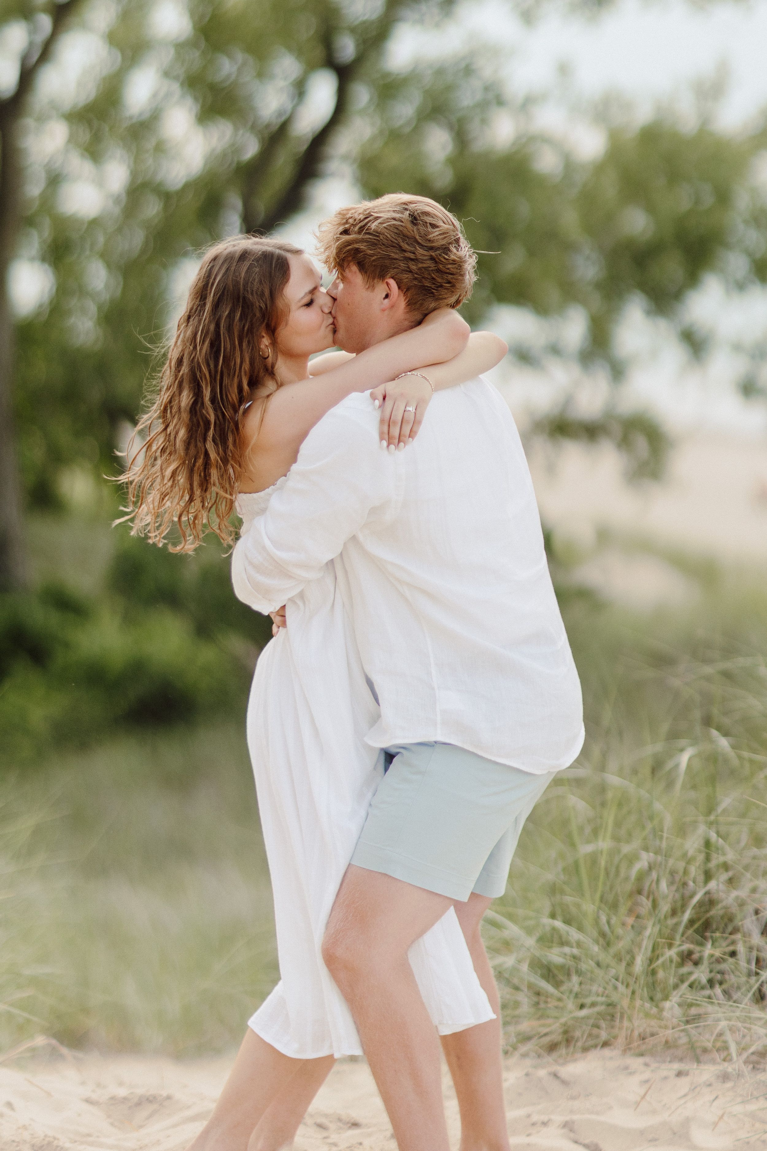 A tender moment between a couple wearing white and blue on a beach with dunes.