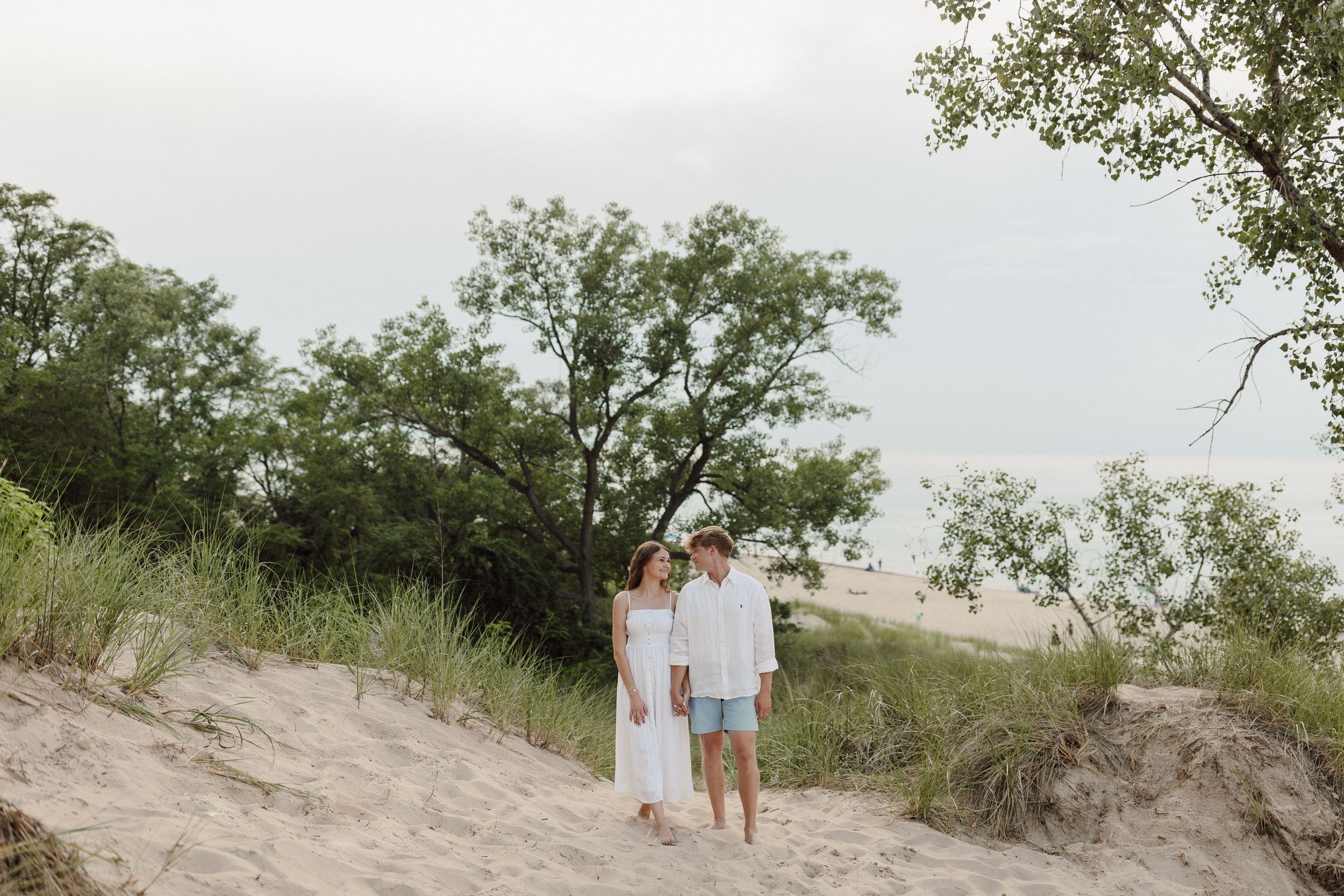 Couple in white outfits walking together along scenic beach dunes.