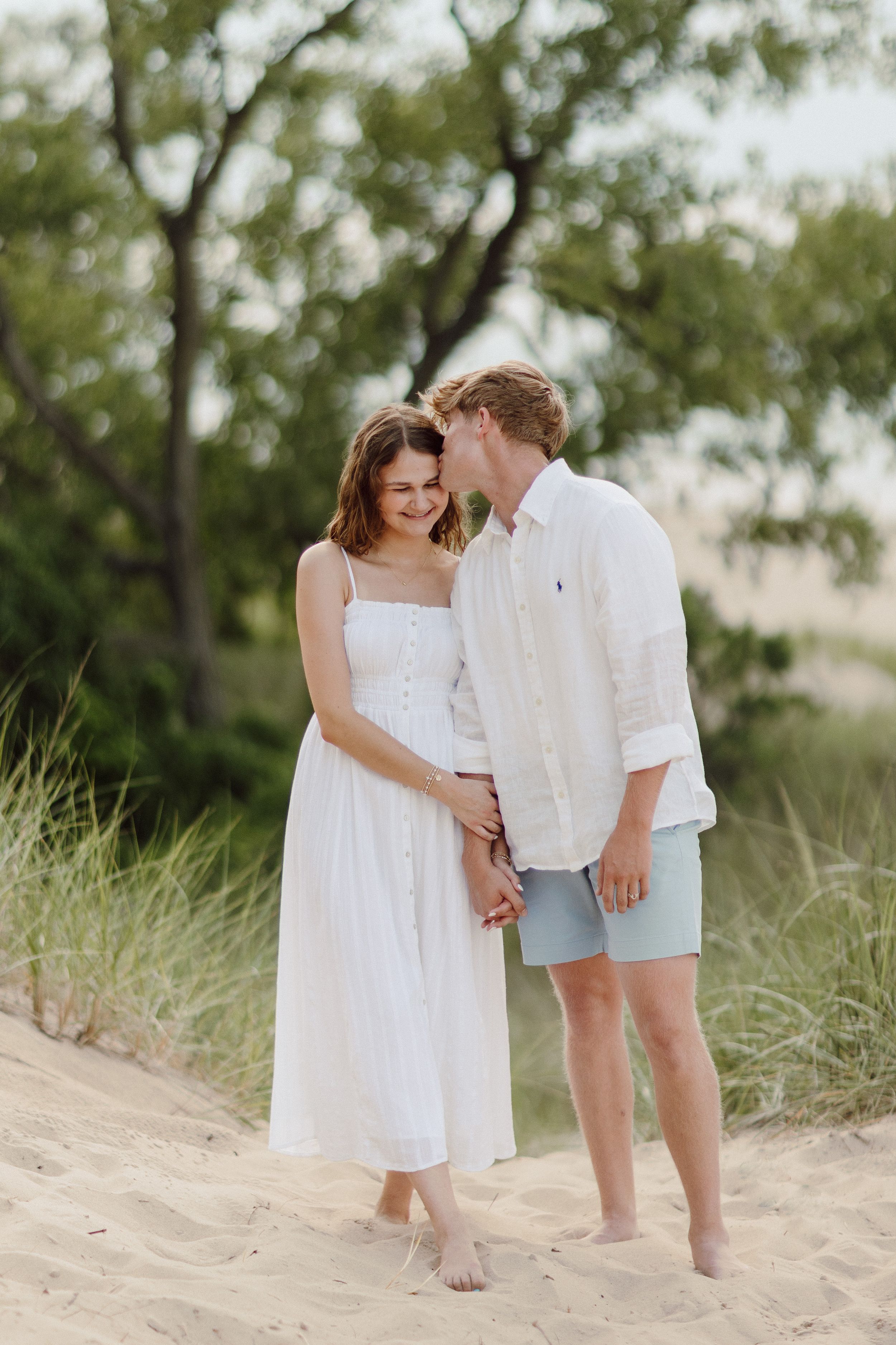 Couple in matching white outfits standing barefoot in beach sand.