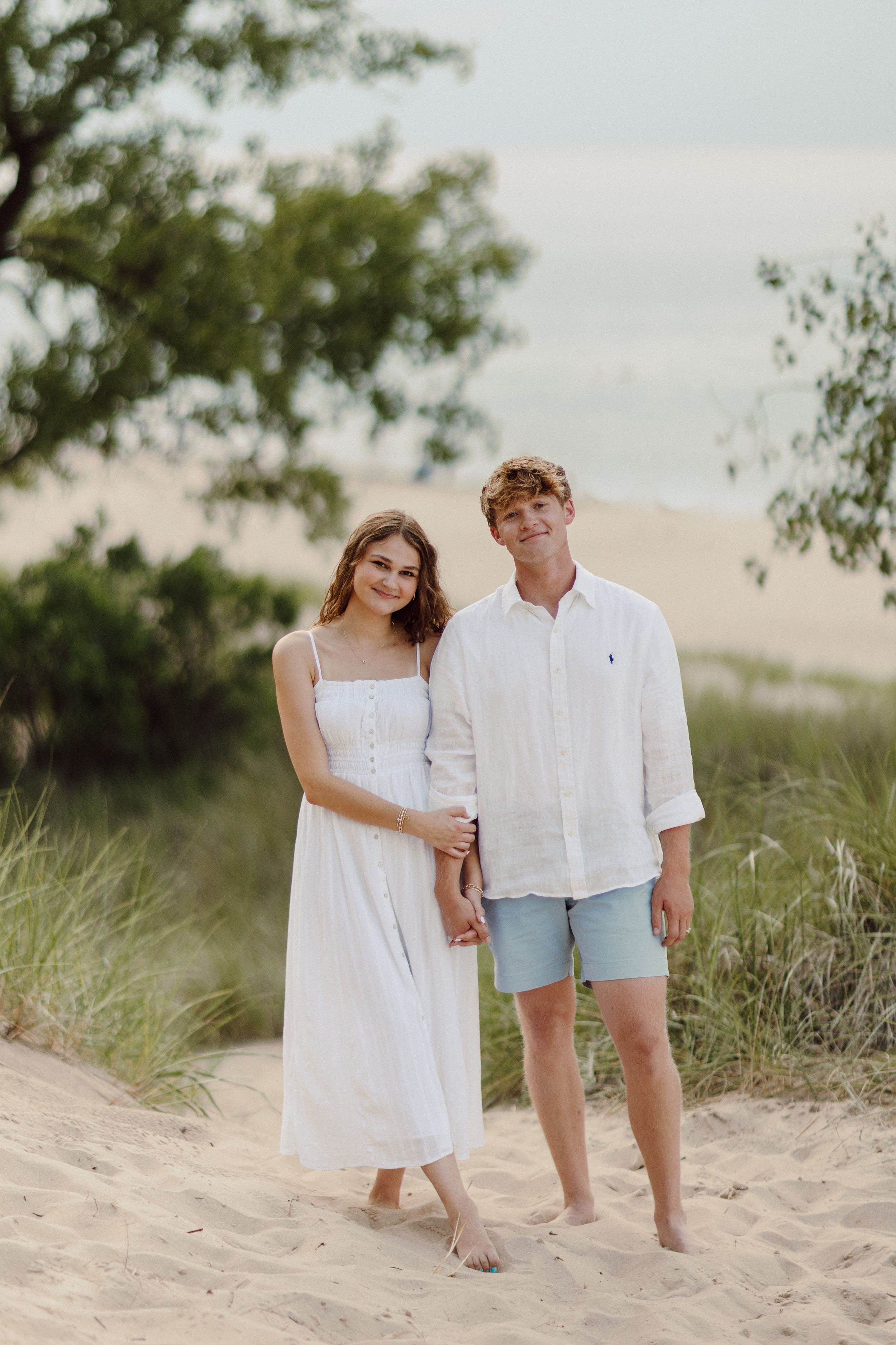 Couple in white summer outfits walking barefoot through beach sand.