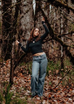 Someone in a black crop top and wide-leg jeans poses gracefully among bare trees in a forest setting.