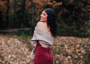Woman in burgundy dress and beige sweater poses playfully among autumn leaves.