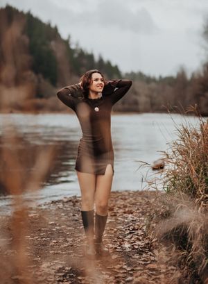 Woman in dark bodysuit and boots poses by riverbank on cloudy day.