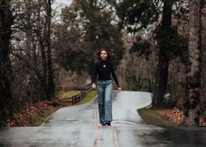 A person in casual attire walks down the center of a wet, winding forest road on an autumn day.