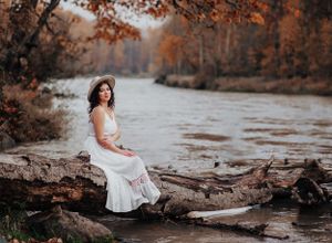 Serene river scene with white-dressed figure seated on fallen log among autumn leaves.