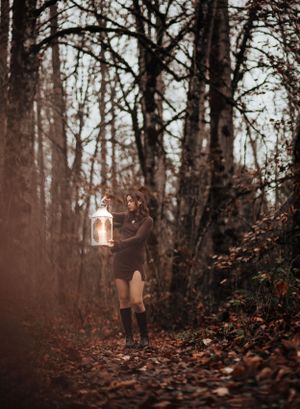 A dreamy autumn forest scene with someone in a brown dress holding a glowing lantern among fallen leaves.