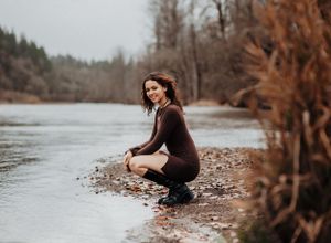 Figure in dark outfit crouches by river's edge surrounded by autumn scenery.