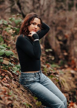 A casual portrait against mossy trees showing someone in a black top and light denim jeans.