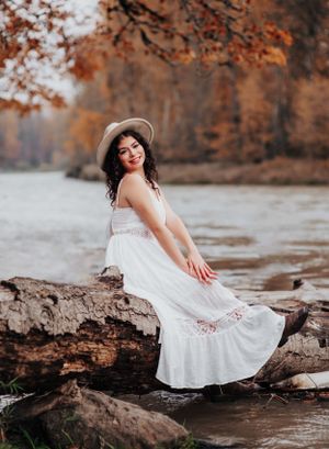 A white lace dress and hat create a dreamy autumn scene by the water with fallen leaves and rocks.