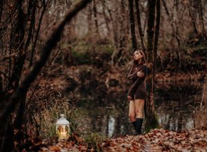 A person in a brown dress poses in a dark forest with a lantern on fallen autumn leaves.