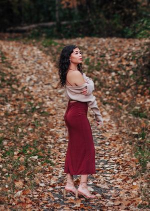 Woman in maroon dress and cream sweater dances amidst fallen autumn leaves on forest floor.