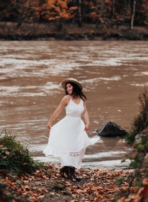 White dress billows in breeze as model poses near muddy river with autumn leaves.