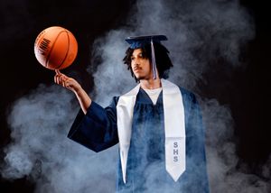 A graduating student poses with a basketball in a dramatic smoky studio setting wearing a cap, gown and graduation stole.