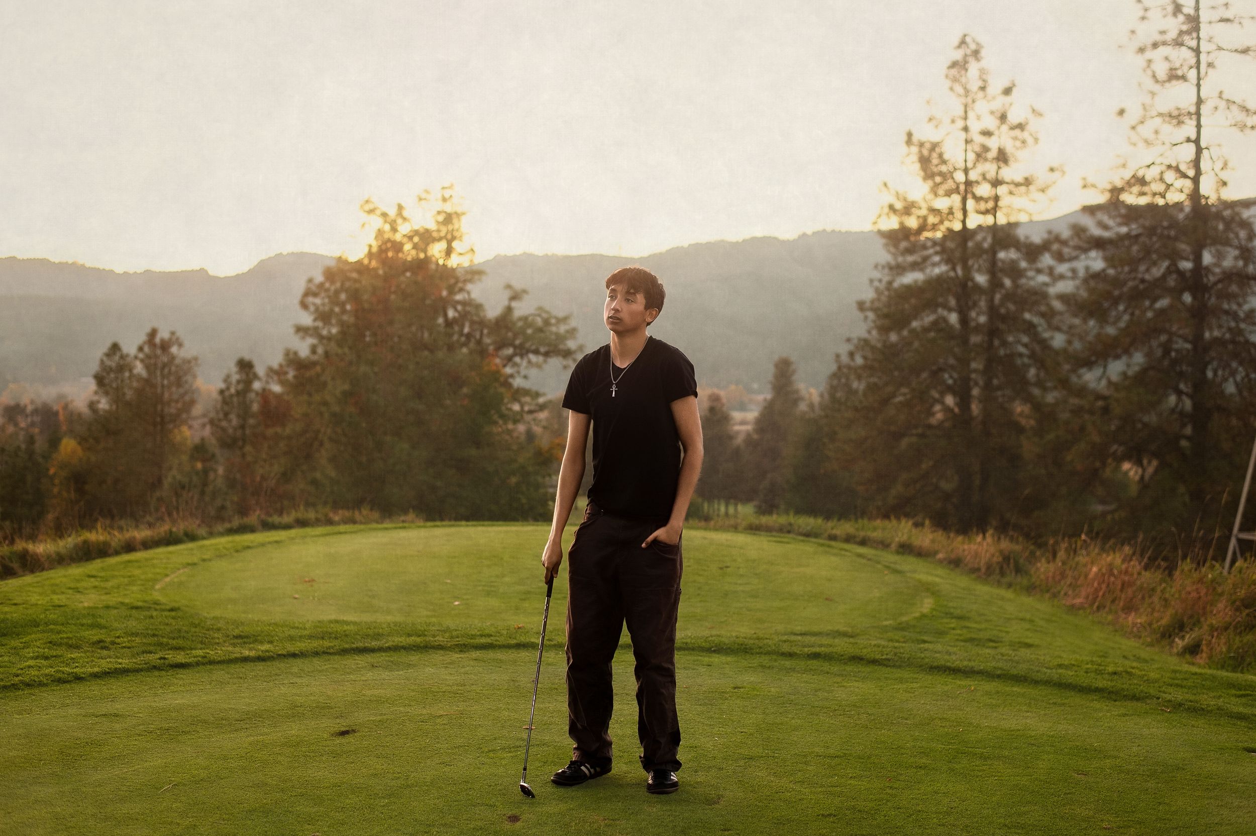 A person stands on a lush green golf course with mountains and trees in the background during golden hour lighting.