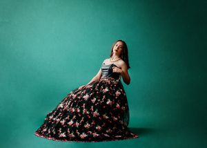 Fashion portrait of a model in a floral black ball gown sitting gracefully against a teal studio backdrop.