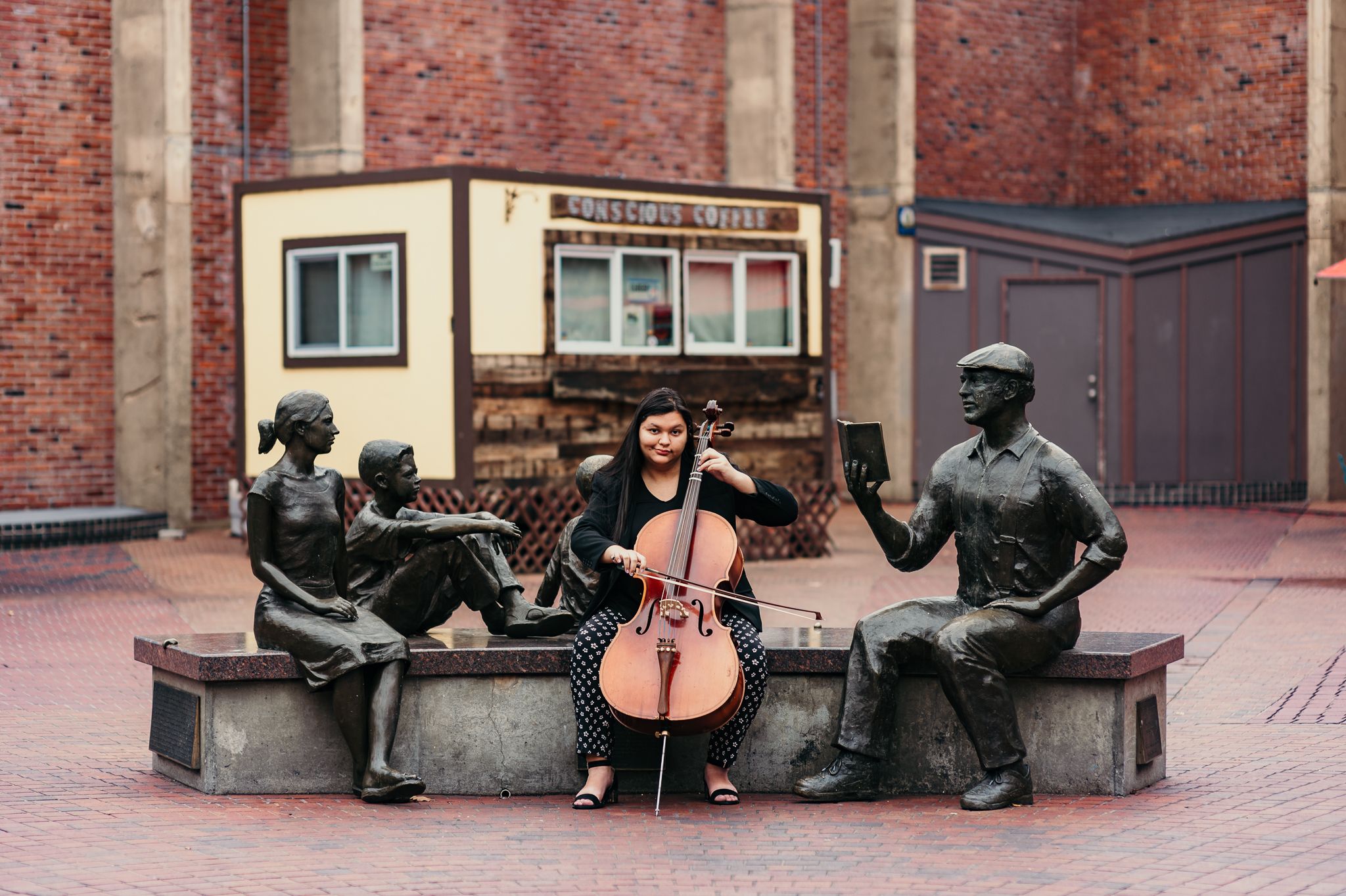Musician with cello seated between bronze statues on urban bench installation.