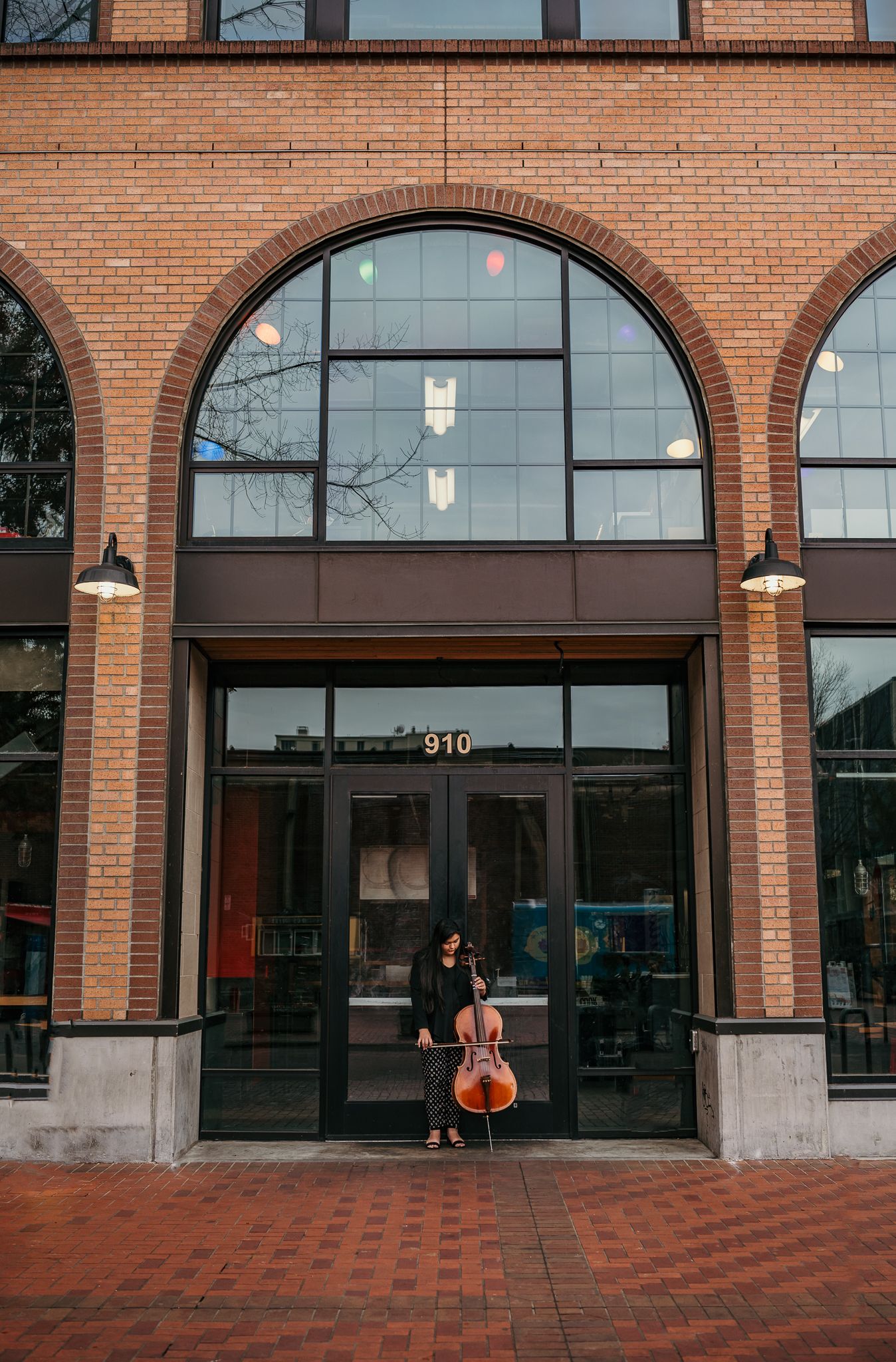 Musician with cello poses under arched brick entrance of modern building.