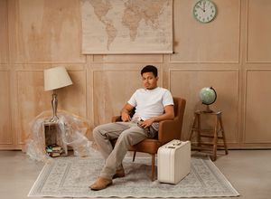 Person sitting in vintage armchair with world map, globe, and suitcase in warm-toned room interior.