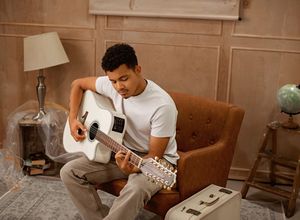 A person in a white t-shirt plays acoustic guitar while sitting in a brown armchair next to a table lamp and globe.