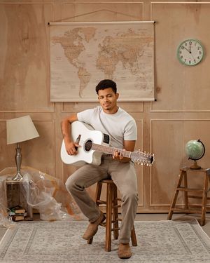 A musician in casual attire sits on a wooden stool playing acoustic guitar in front of a world map backdrop.